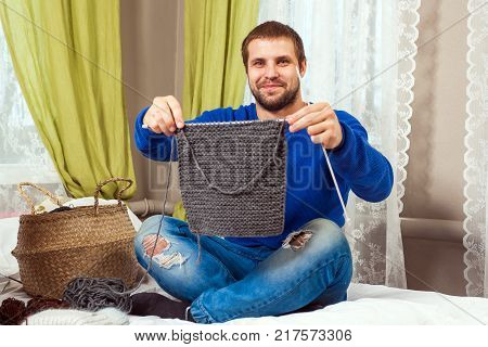 A young dark haired man in a blue sweater and jeans shows how he tied a piece of a gray sweater with knitting needles from natural strings and sits on a bed in a home setting
