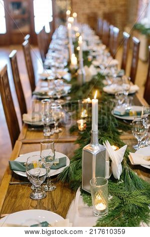 holidays, interior, amusement concept. adorable christmas decor of the long table in restaurant, created by candles in hamdmade holders of silver colour and branches of some fur tree