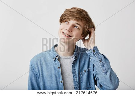 Portrait of pensive fair-haired dreamy hipster guy imagines something in his mind, looks aside, smiles, being deep in thoughts, scratching head. Attractive hipster guy dreams about vacation or weekends alone