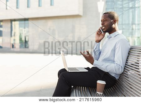 Happy black businessman working on laptop. African-american insuranse agent checking email on computer, talking on mobile, sitting on the bench outdoors near modern office center