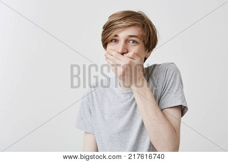 Frustrated puzzled shocked male with fair hair and boue eyes keeps hand on mouth, being surprised to make big mistake or fail exam. Terrified amazed fearful caucasian young man in astonishment and confusion