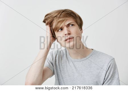 Headshot of clueless perplexed young caucasian man in grey t-shirt looking aside with confused and puzzled expression, scratching head, trying to remember something important, forgot about his girlfriend's birthday