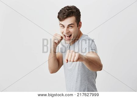 Positive confident muscular young sportsman with dark hair and blue eyes dressed in gray shirt smiling broadly, holding fists in front, ready for new challenges, demonstrating his strength