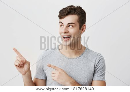 Horizontal portrait of caucasian handsome man having broad smile wearing gray t-shirt posing against studio wall pointing with index fingers at white copy space for advertisment or promotional content.