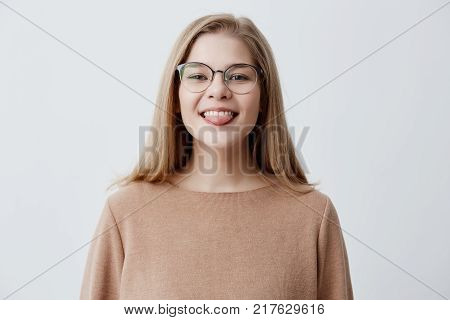 Horizontal portrait of optimistic funny girl with blonde straight hair wearing brown sweater and glasses showing her tongue while posing against gray studio background. Emotional positive young female making faces