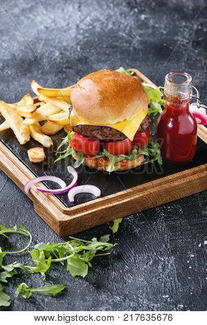 Homemade traditional hamburger with beef, tomato, cheese, aragula, served on wooden slate serving board with french fries and ketchup sauce on dark texture background.