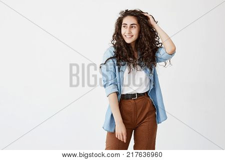Horizontal portrait of beautiful good looking girl with appealing appearance smiles happily, wears her long wavy hair loose, has good mood after spending weekends with friends, glad and pleased to pose at camera