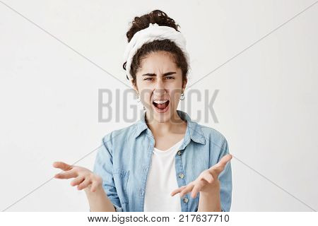 Close up isolated portrait of young dark-haired annoyed angry woman in do-rag and denim shirt holding hands in furious gesture. Young female in white T-shirt expresses negative emotions and feelings