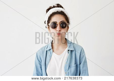 Funny female model with dark hair in bun pouts lips, in denim shirt and roung sunglasses, makes grimace as stands in studio. Joyful pretty woman has fun indoors, blows cheeks, foolishes at camera