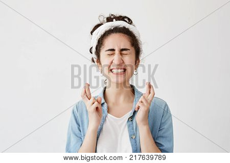 Glad supertitious young brunette girl in do-rag and denim shirt with closed eyes crosses fingers, clenches teeth, prays before important exam, wishes luck, hopes for victory and success. Feeling hopeful. Waiting for miracle