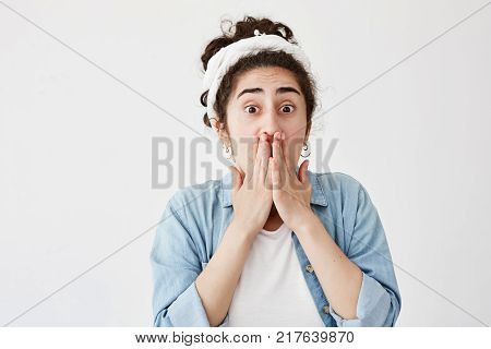 Shocked scared young female with dark hair wearing do-rag and denim shirt covering mouth with both hands keeping secret. Girl doesn't want to spread confidential information, poses with popped eyes