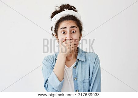 Portrait of shocked scared young brunette wearing do-rag and denim shirt covering mouth with hand keeping a secret. Dark-haired young female doesn't want to spread rumors or some confidential information, poses with popped eyes