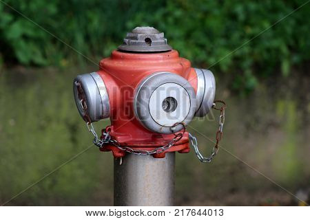 Metal red and grey fire hydrant top with four caps connected with small partially rusted chains on green leaves and stone background