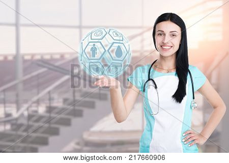 Woman doctor isolated on a clinic background, holding a graphic sphere with medical icons. Concept: medicine, innovative medical technology, healthcare.