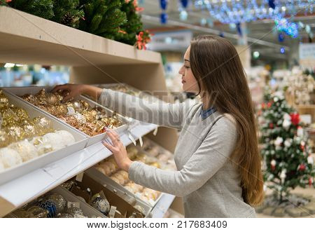 Beautiful happy customer girl choosing gifts for Christmas and New Year. She is taking Christmas tree decorations. Big supermarket sale during New year