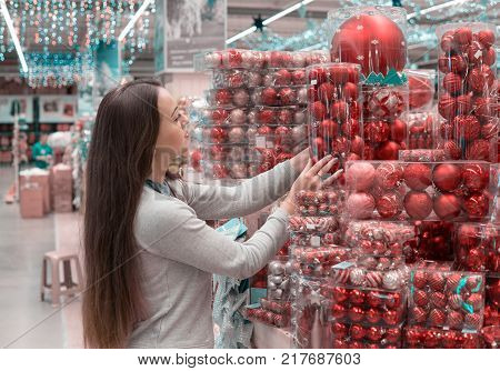 Cheerful happy customer girl choosing gifts for Christmas and New Year. She is taking red Christmas tree decoration balls. difficult decision. Various choice