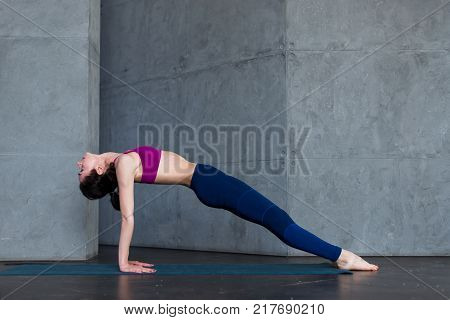 Pretty thin woman in sports bra and leggings doing yoga upward facing plank pose, purvottanasana, working out indoors.