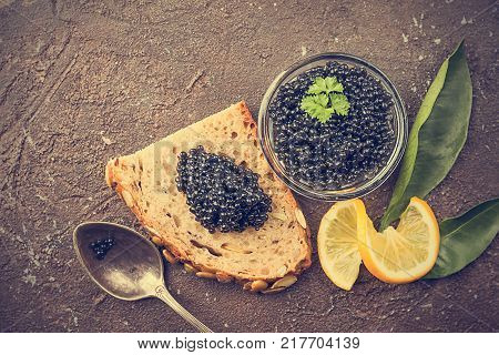 Black caviar in a glass bowl with lemon close up over dark background, top view