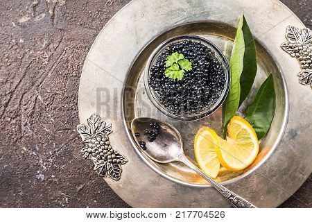 Black caviar in a glass bowl with lemon close up over dark background, top view