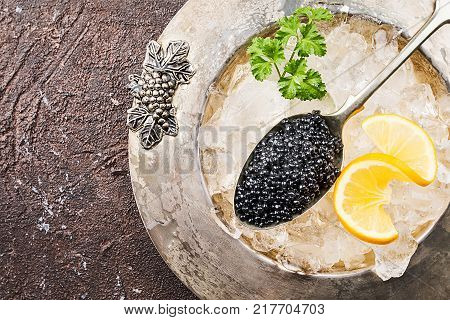 Black caviar in a spoon on ice with lemon close up over dark background, top view