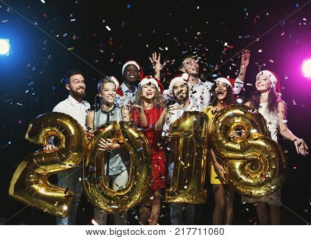New 2018 year is coming. Group of cheerful young people in Santa hats holding gold colored number balloons, showered with confetti. Christmas greeting card mokup, celebration concept
