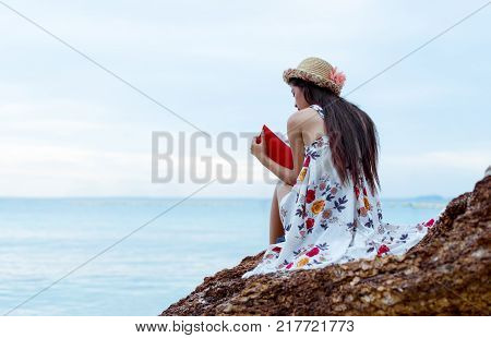 Asian woman reading book alone at beach lonely with wearing white dress in summer vacation relaxing in tropical sea