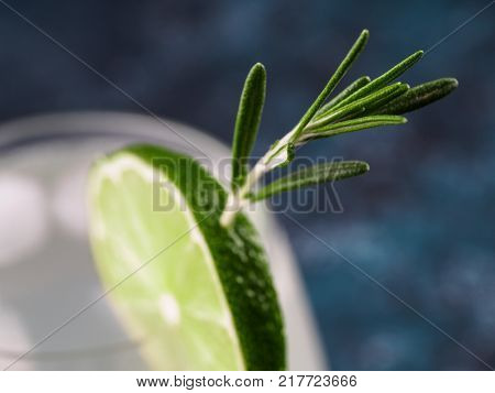 Glass of alcoholic coctail with fresh rosemary and lime on blue background. Close up. Shallow DOF