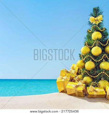 Christmas tree with golden balls and fancy gift boxes with bows at tropical sandy beach against ocean. New Year holidays in hot countries concept square greeting card with space for your text