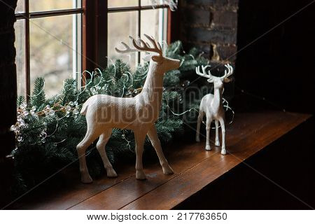 Christmas decoration with pine tree branches and white clay deers standing on wooden window sill. New year decor