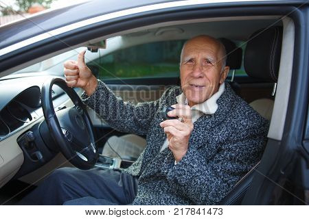 The old gray-haired man is sitting in the car behind the wheel, holding in his hand a small action packed camera and showing a thumbs-up gesture. Outdoors on the street.