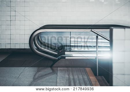 Side view of modern glass and chrome escalator in urban underground with white tiles in background; city contemporary moving staircase with stairs next to it following down into subway