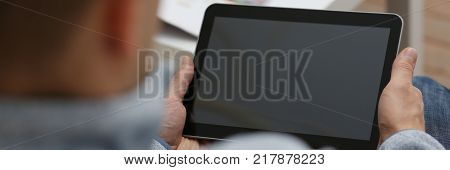 Male hand holds tablet   in home setting while sitting on the couch engaged an internet surfing using application to press a finger on the display leisure listering music concept closeup.