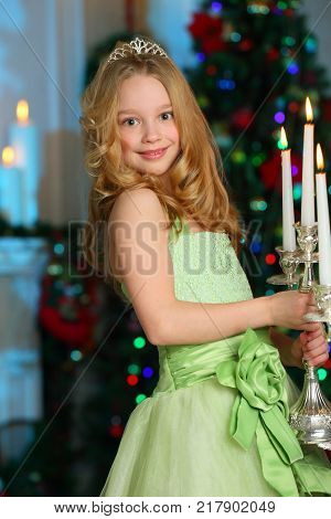 Beautiful charming pretty blond child-girl in a festive green dress with luminous candles on the background of a New Year tree.