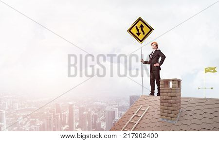 Young determined businessman standing on house roof and showing yellow roadsign. Mixed media