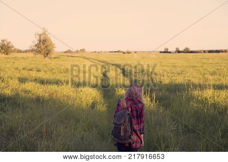 girl in plaid shirt with a backpack on shoulders walking down country road in the middle of a field with tall grass