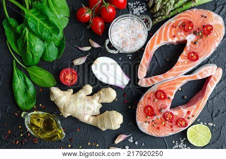 Two fresh raw salmon steaks with asparagus spinach garlic tomatoes ginger pepper corns salt chili onion lemon and olive oil on black background. Healthy food diet concept. Top view.