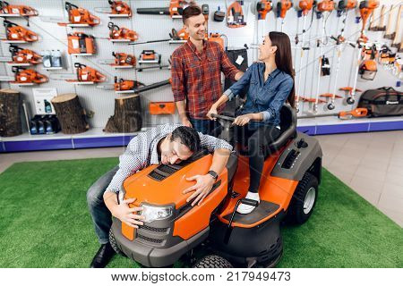 A consultant in a garden tools store shows a guy and a girl a lawn mower. There are many tools for gardening here. A young family came to choose their equipment.