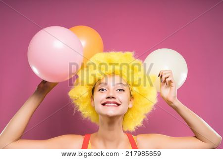 funny laughing young girl with a clownish yellow wig on her head, holding multicolored balloons in her hands