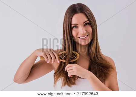 Hair care and treatment concept. Portrait of young brunette woman combing her hair and smiling while isolated on white background. Beautiful girl wrapped in towel with comb in hand