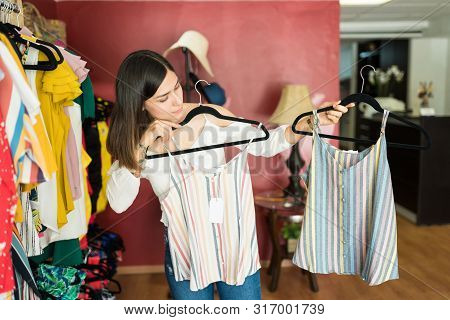 Female Customer Comparing Striped Tops Hanging From Coathangers In Shopping Mall