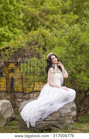 Sensual Caucasian Bride With Diadem Sitting On Stone In Flowers Garden Outdoors. Wearing Beads Neckl