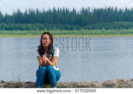 Surprised Yakut Asian Young Girl Sitting On A Stone Shore With Folded Hands In The Background Of The