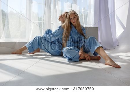 Two Young Pretty Twin Girls In Identical Blue Suits With Long Blond Hair Sitting On White Cyclorama 