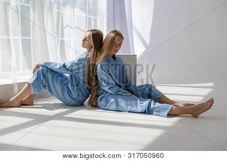 Two Young Pretty Twin Girls In Identical Blue Suits With Long Blond Hair Sitting On White Cyclorama 