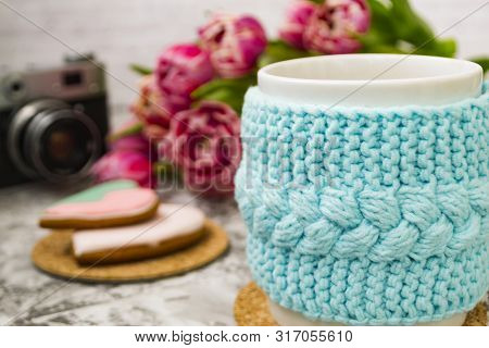 A White Cup Of Tea In A Knitted Blue Case With An Old Camera And Pink Flowers Tulips And Sweets, Gin