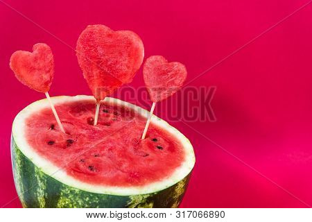 Half Ripe Juicy Watermelon Decorated With Decorative Hearts On A Wooden Stick Isolated On A Red Back