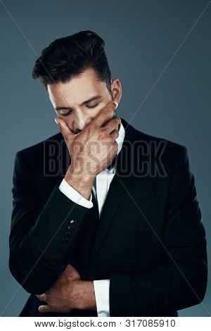 Tired Young Man In Bow Tie Covering Mouth With Hand While Standing Against Grey Background