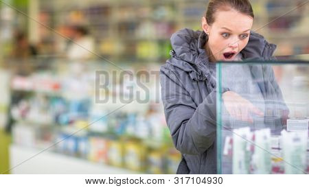 Young woman looking for the right pills/her prescription in a modern pharmacy -disturbed by the exorbitant prices of the meds(shallow DOF; color toned image)