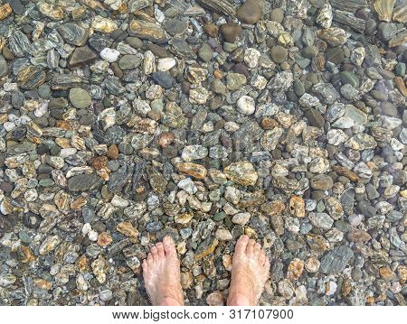A Pair Of Feet Standing On Colourful Pebbels In The Shallow Waters Of A Crystal Clear Lake In New Ze