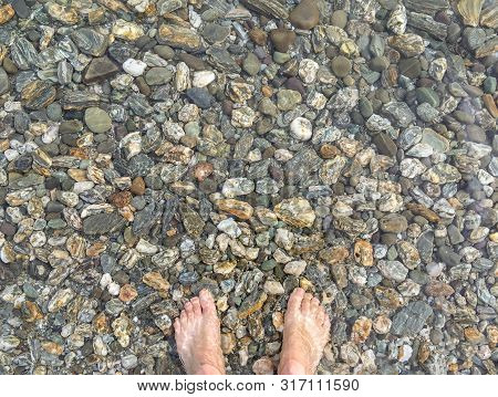 A Pair Of Feet Standing On Colourful Pebbels In The Shallow Waters Of A Crystal Clear Lake In New Ze